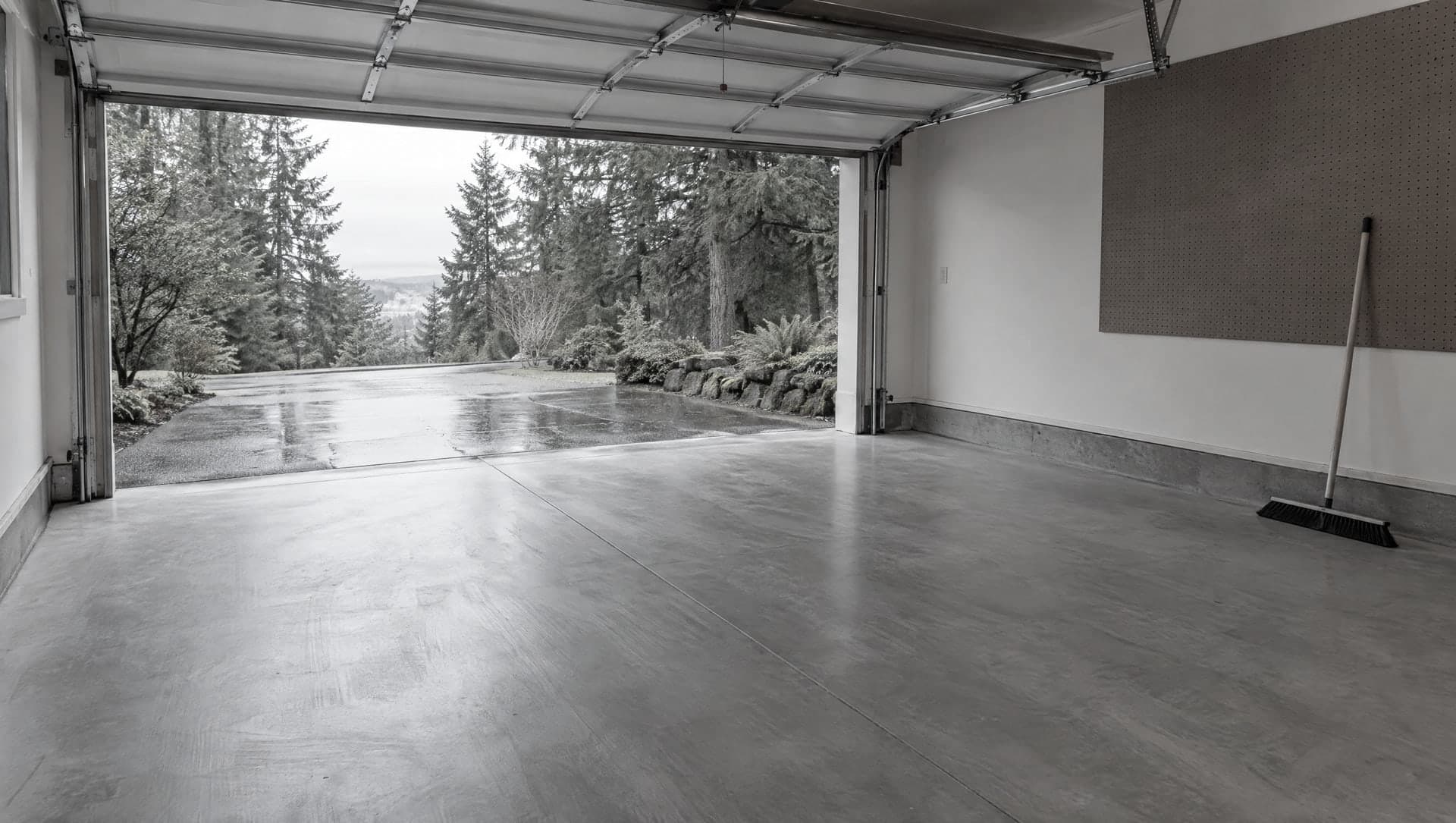 An empty, swept garage with its door open onto a wet driveway and a broom against the wall.